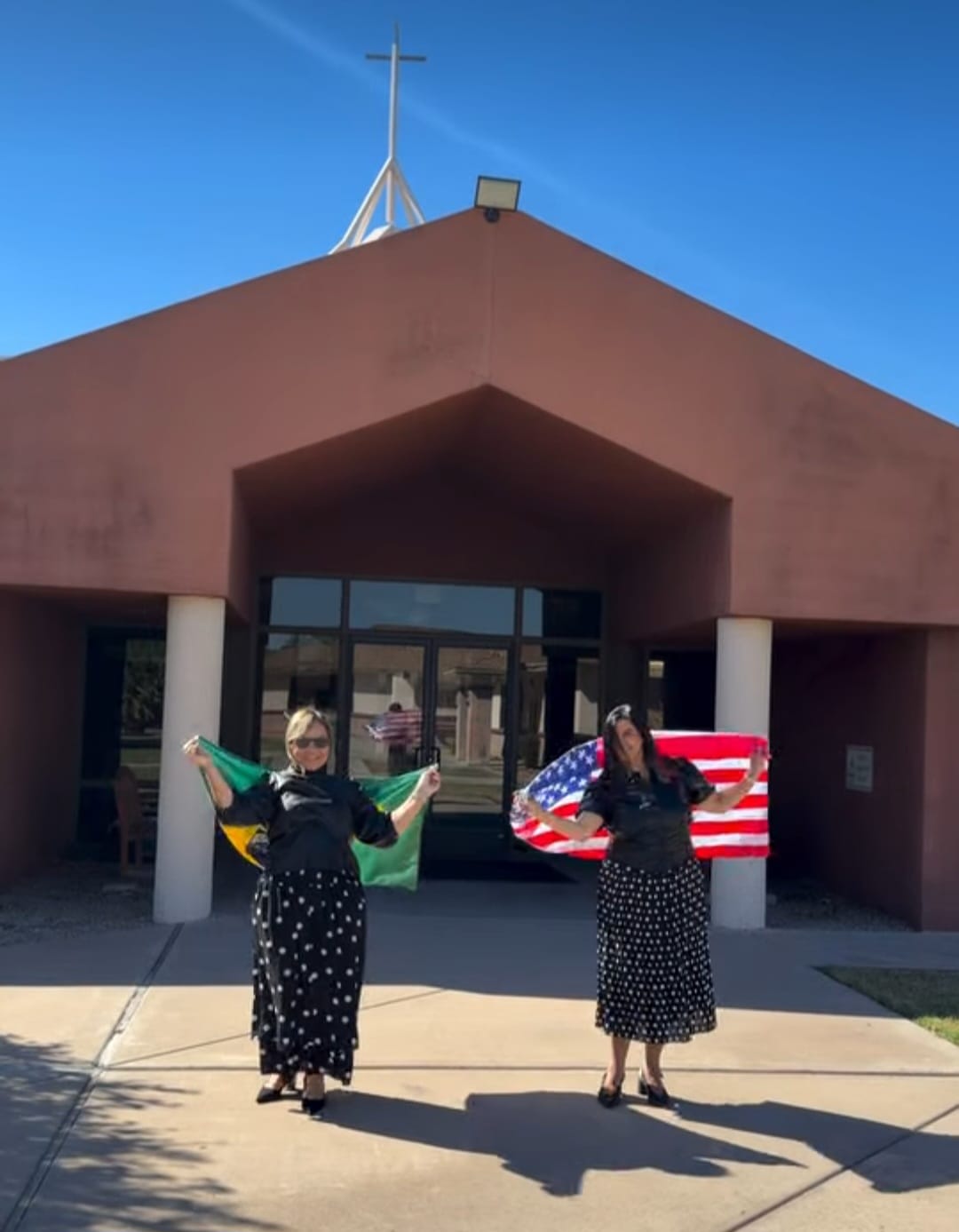Sandra Martins e outra mulher segurando as bandeiras do Brasil e dos Estados Unidos em frente a uma igreja
